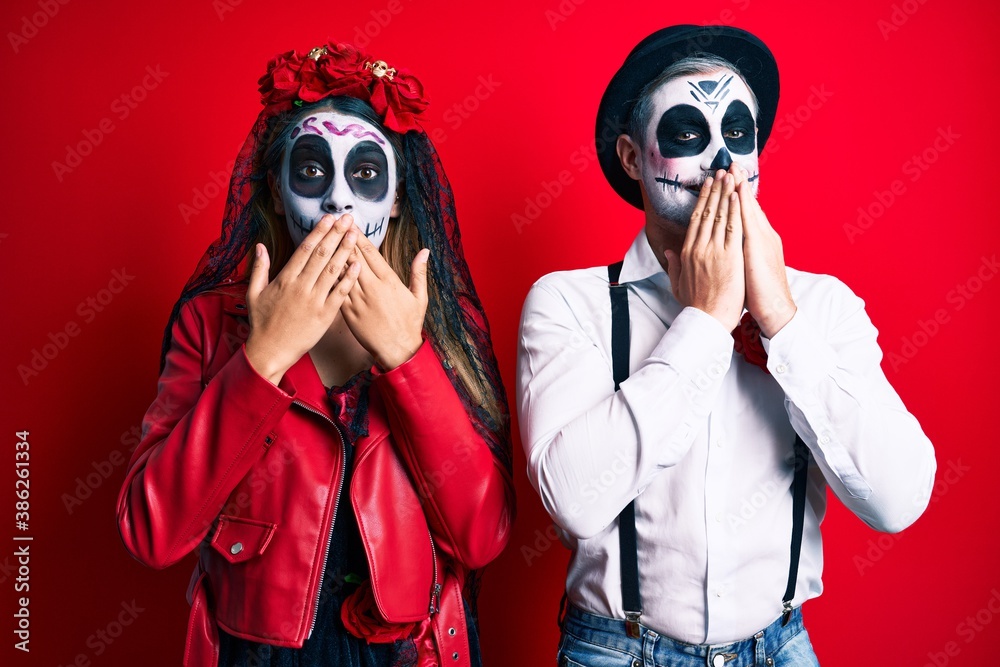 Couple wearing day of the dead costume over red laughing and embarrassed giggle covering mouth with hands, gossip and scandal concept Couple wearing day of the dead costume over red laughing and embarrassed giggle covering mouth with hands, gossip and scandal concept