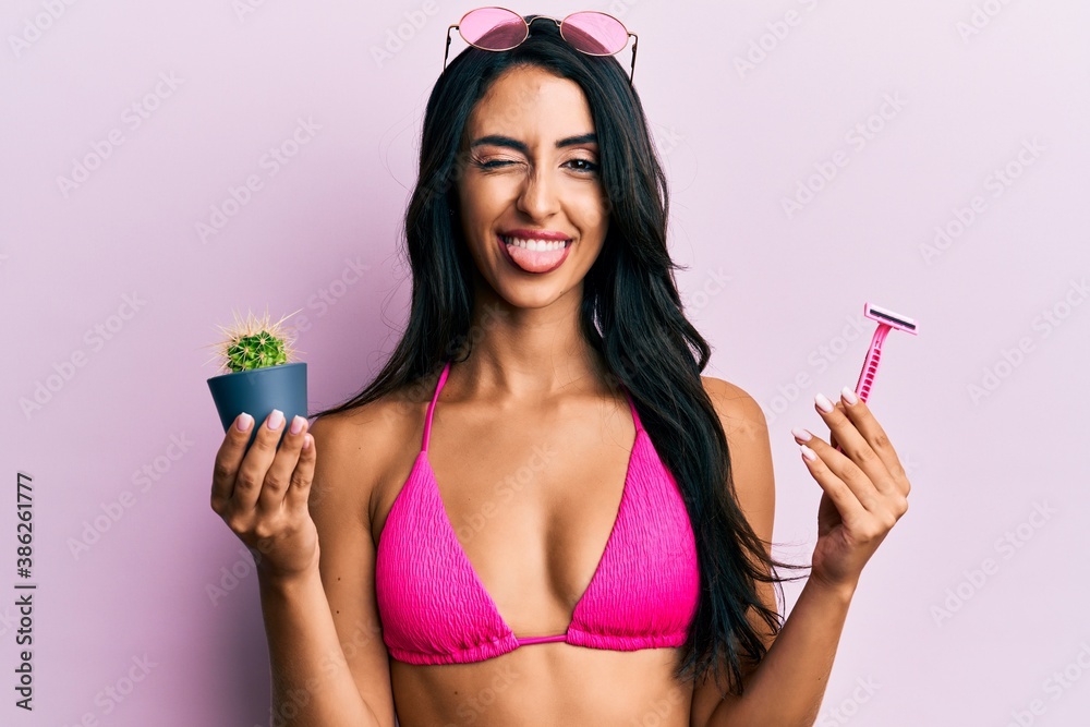 Beautiful hispanic woman wearing bikini holding cactus and razor sticking tongue out happy with funny expression. Beautiful hispanic woman wearing bikini holding cactus and razor sticking tongue out happy with funny expression.