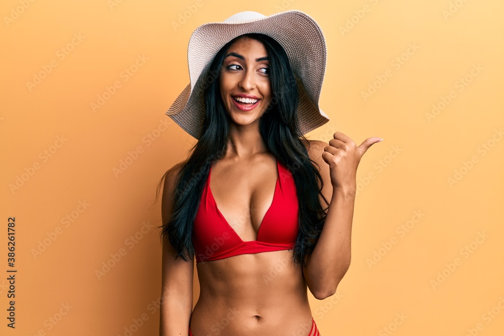 Beautiful hispanic woman wearing bikini and summer hat pointing thumb up to the side smiling happy with open mouth Beautiful hispanic woman wearing bikini and summer hat pointing thumb up to the side smiling happy with open mouth