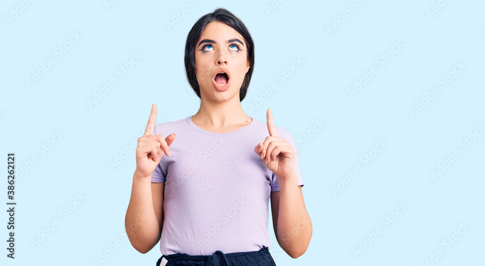 Brunette teenager girl wearing casual clothes amazed and surprised looking up and pointing with fingers and raised arms. Brunette teenager girl wearing casual clothes amazed and surprised looking up and pointing with fingers and raised arms.