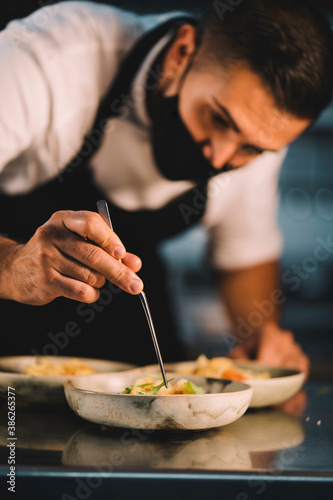 Close-up of a male chef with mask decorating food in ceramic dishes over stainless steel worktop in restaurant kitchen during coronavirus pandemic.
