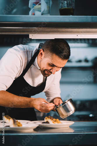 Portrait of a male chef decorating food with spoon in ceramic dish over stainless steel worktop in restaurant kitchen. photo