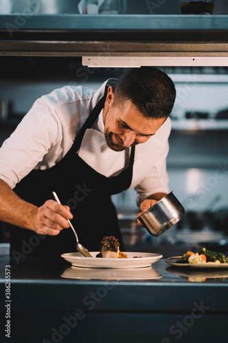 Portrait of a male chef decorating food with spoon in ceramic dish over stainless steel worktop in restaurant kitchen.