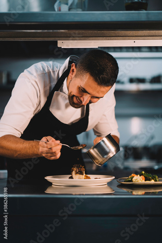 Portrait of a male chef decorating food with spoon in ceramic dish over stainless steel worktop in restaurant kitchen. © Henko Studio