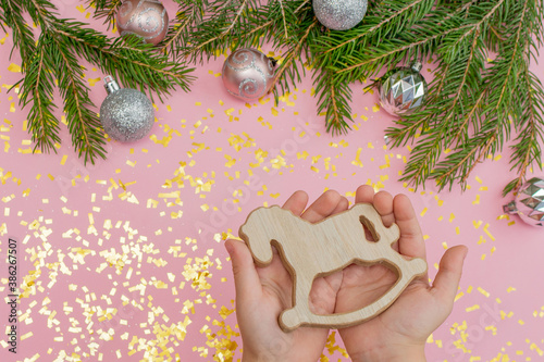 in children's hands - a wooden Christmas tree toy in the form of a rocking horse, on a pink background with fir branches, Christmas balls. Close-up, pastel background with golden confetti, soft focus. photo