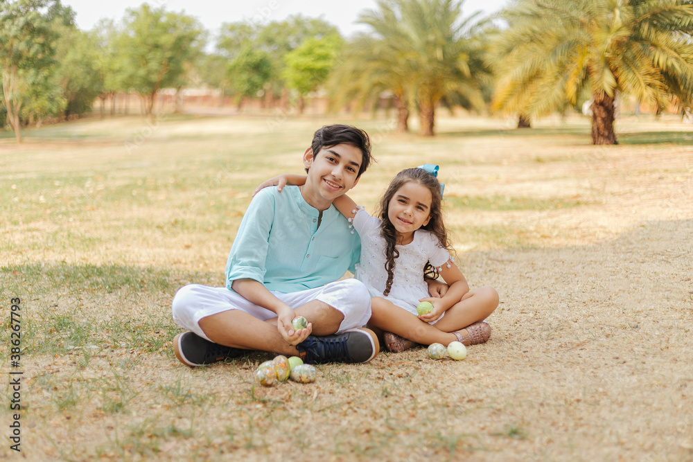 Latin brothers celebrating Easter. Curly girl and guy sitting on the grass in a park hugging and holding easter eggs