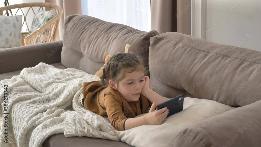 Pretty little child with bows covered with knitted plaid watches cartoons on cellphone on comfortable sofa in living room Pretty little child with bows covered with knitted plaid watches cartoons on cellphone on comfortable sofa in living room