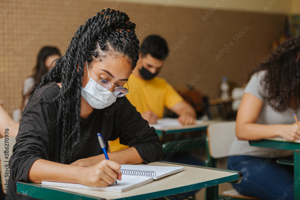 Naklejka premium Latin students in the classroom. female student with twisted hair wearing mask and writing in notebook with a pen. Covid-19. Pandemic.