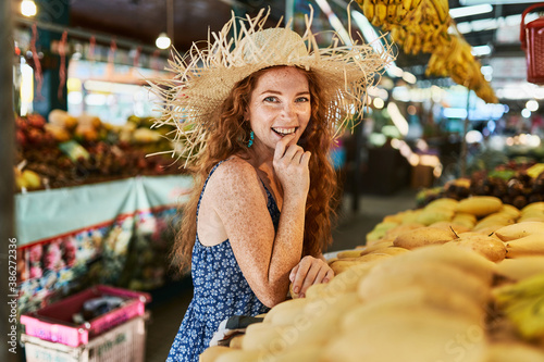 A young smilling  redhead girl in hat holding near yellow mango in her hand at the marketplace in Thailand. Copy space © Nikolay