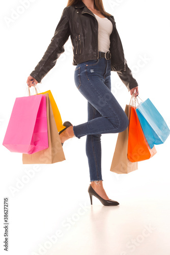 Girl with shopping after Black Friday stands with her leg raised. A shopaholic girl in a leather jacket stands on a white background with packages in hands after sale.