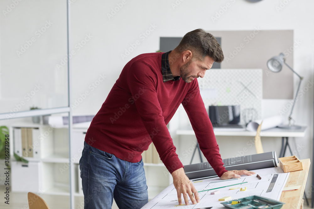 Side view portrait of bearded architect looking at blueprints while leaning on drawing table at workplace, copy space