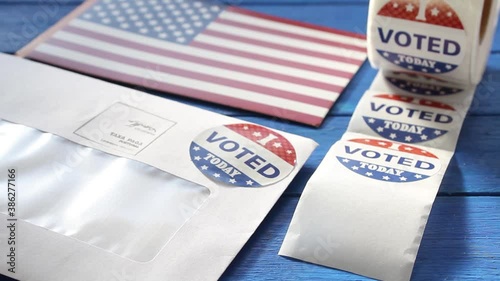 Woman's hand taking a I Voted today sticker from a roll on table with envelope and USA flag. Vote by mail in US presidential election concept