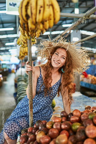 Young smiling redhead girl with freckles in a blue dress and a straw hat sits on the counter with mangoosteens and babanas. Fruit market in Thailand