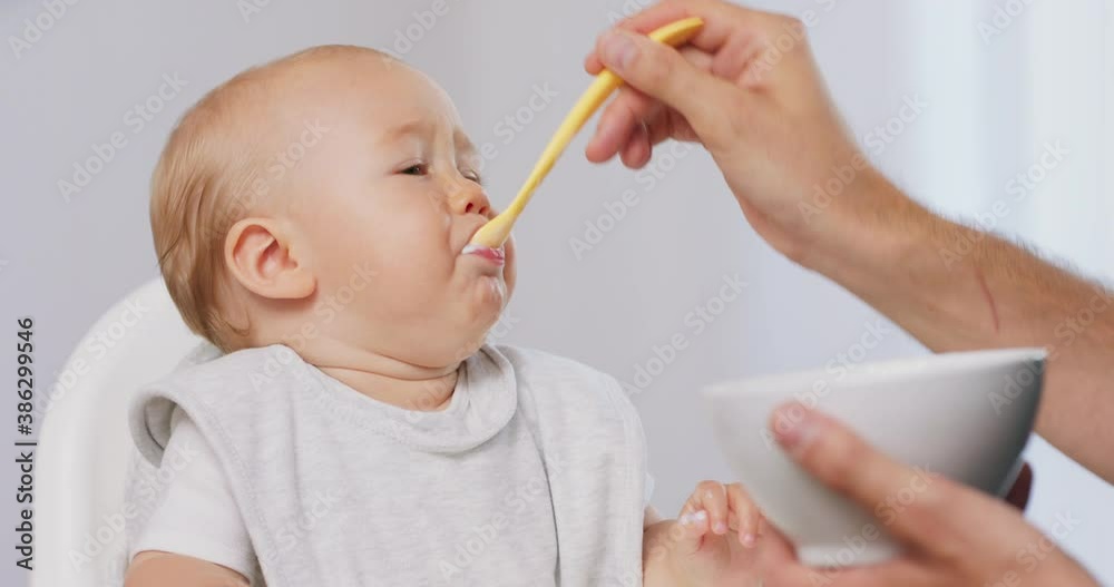 Closeup of young baby in white high baby chair and father's hand with a spoon and bowl, while he is feeding the baby, who eats and makes funny grimaces. White walls on background. Closeup of young baby in white high baby chair and father's hand with a spoon and bowl, while he is feeding the baby, who eats and makes funny grimaces. White walls on background.