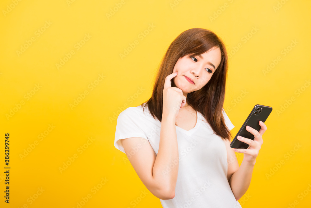 Asian happy portrait beautiful cute young woman teen standing her using holding smart mobile phone and thinking looking to the phone isolated, studio shot yellow background with copy space Asian happy portrait beautiful cute young woman teen standing her using holding smart mobile phone and thinking looking to the phone isolated, studio shot yellow background with copy space