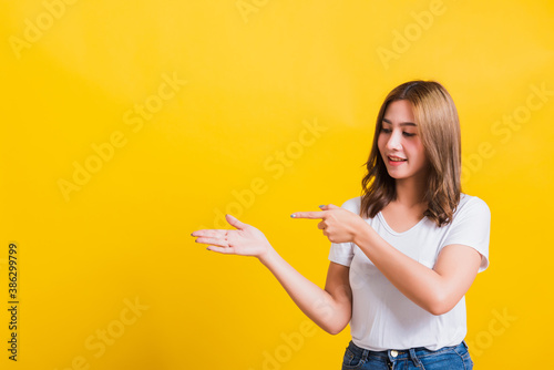 Asian Thai happy portrait beautiful young woman standing to hold something on palm away side and point the finger to it looking to side  studio shot isolated on yellow background with copy space