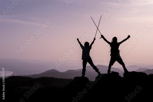 Happy winners reaching life goal - success people at summit. Business achievement concept. Two person couple together arms up in the air of happiness with accomplishment in the clouds at sunset.