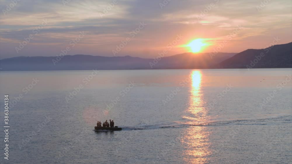 Dinghy with people on it moving from right to left at sunset, with mountains in the background and orange sky, shot near Portofino (Italy) Dinghy with people on it moving from right to left at sunset, with mountains in the background and orange sky, shot near Portofino (Italy)