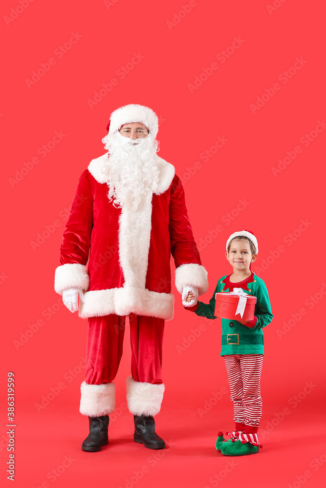 Fototapeta premium Cute little elf with Santa Claus and Christmas gift on color background