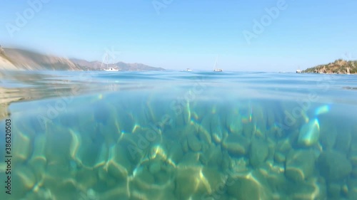 Slow motion footage of submersion underwater in crystal clear turquoise sea on a stony  rocky beach with anchored boats and hills in the background