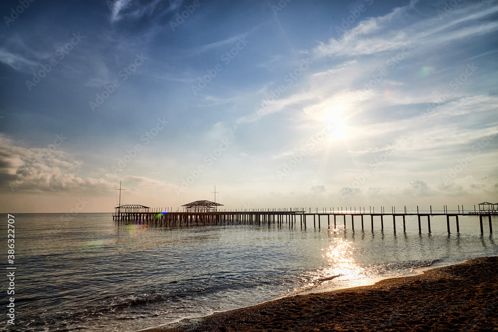 View from sand beach to water of sea, waves and pier in a nice day or evening with blues sky, bright sun and white clouds. The concept of a holiday on the sea or ocean in the South.