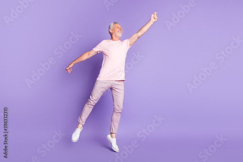 Full length profile photo of old man tiptoe raise hand look empty space wear pink t-shirt pants sneakers isolated purple color background