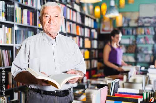 Old positive belgian male is choosing book for reading in free time in bookstore.