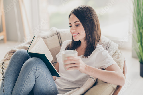 Photo of pretty young girl sit chair read interesting book hold tea cup wear denim jeans in house indoors photo