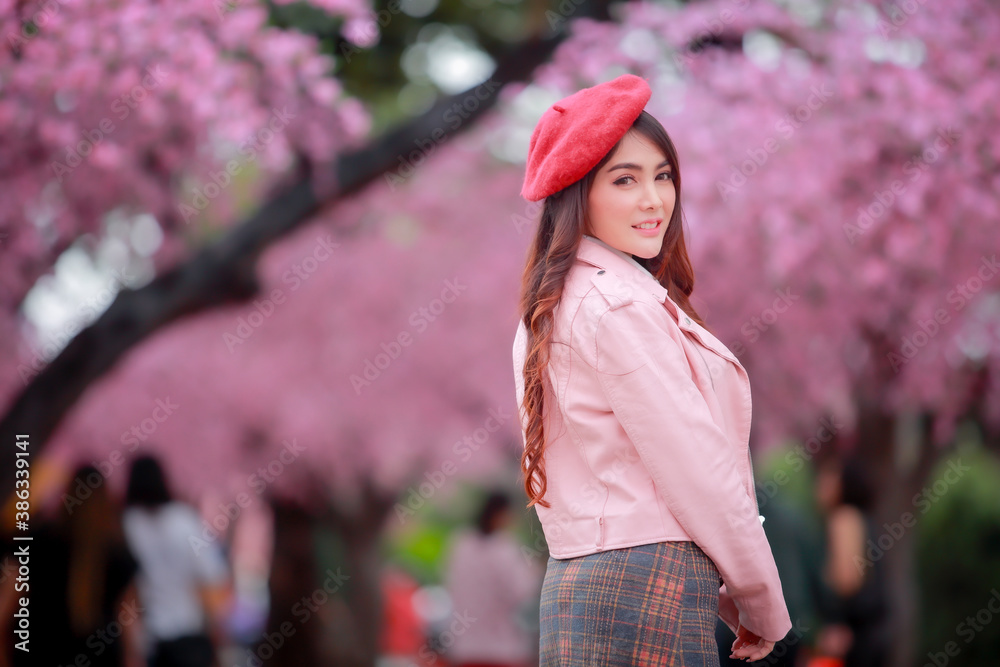 A traveler hipster woman sightseeing wear a red hat and a smooth leather dress with beautiful sakura cherry blossoms tree full blooming in pink color in the park on a spring day.