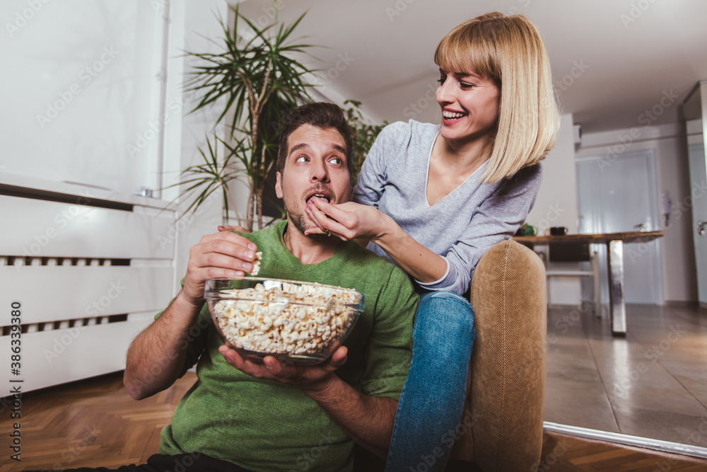 Young couple sitting together on a sofa at home watching television, joyfully smiling eating pop corn enjoying a night in together.