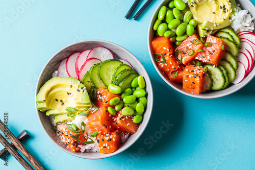 Poke bowl with salmon, rice, avocado, edamame beans, cucumber and radish in gray bowls, top view. Hawaiian ahi poke bowl, blue background.