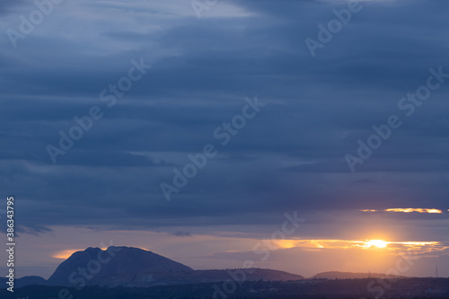 A Panoramic Silhouette of hills in the bottom with dark clouds in the horizon with sun peeping through them
