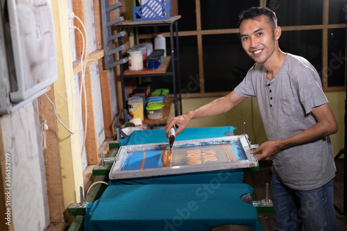 Young male worker smiles when holding squeegee blade and screen printing frames for t shirt screen printing