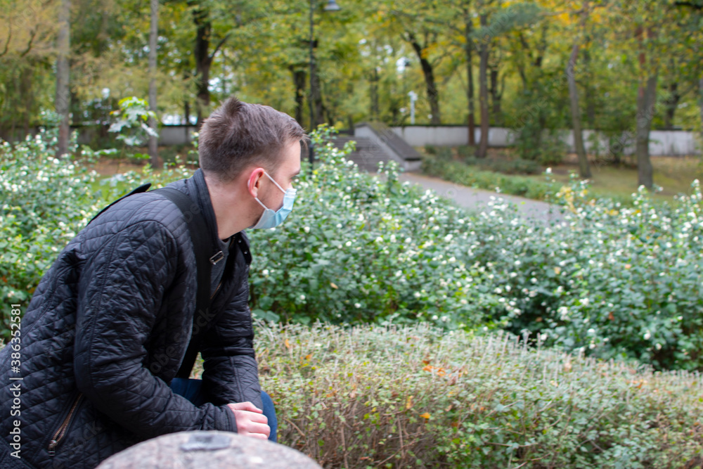 a young adult guy stands in a mask in the park looks to the side thoughtfully, in dark clothes, a green park a young adult guy stands in a mask in the park looks to the side thoughtfully, in dark clothes, a green park