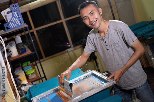 Young Asian male worker smiles when preparing screen printing frames for t shirt screen printing with holding squeegee blade