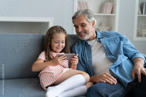 Cute preschooler girl help father show something on smartphone, smart little daughter and dad sit on couch hold mobile phone