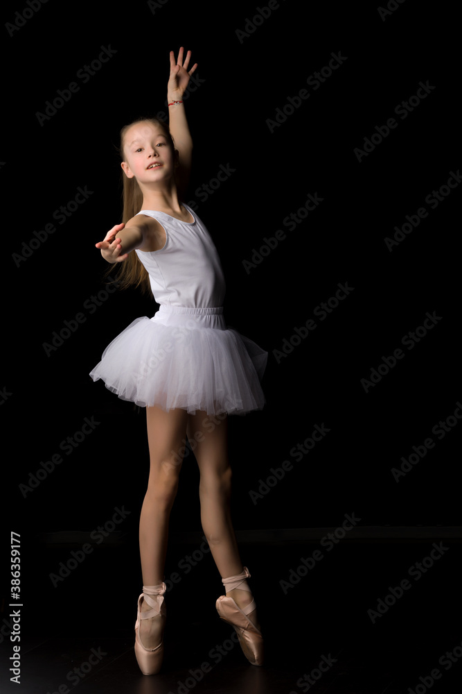 Naklejka premium Cute little girl in a tutu and pointe shoes is dancing in the studio on a black background.