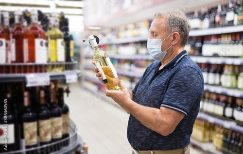 Confident elderly man in protective mask choosing white wine in modern wineshop