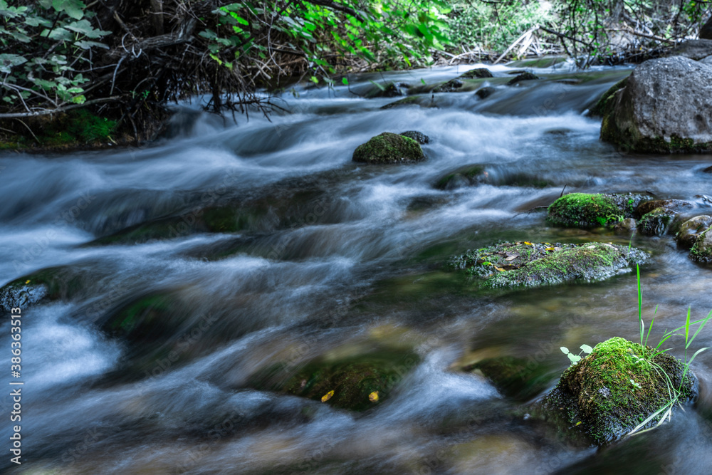 Obraz premium Fast running mountain river with mossy boulders and rocks at cloudy day on long exposure.