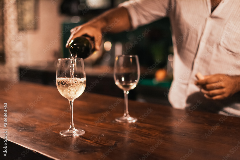 A barman pouring white wine into a glass. Wooden table in a dark bar