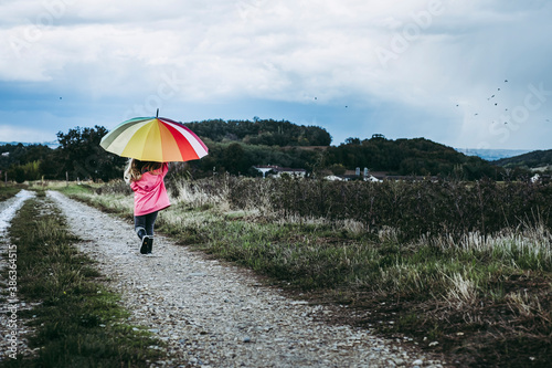 Petite fille avec un parapluie arc en ciel en train de marcher sur un chemin par temps de pluie