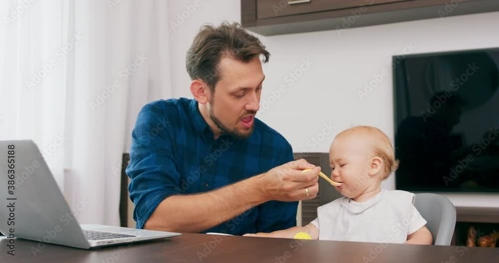 Young bearded father, and a cute baby with bib, in high baby chair, are sitting at the desk with laptop on it. Father feeds baby with bowl and spoon. Baby makes funny faces and grimaces. Slow motion Young bearded father, and a cute baby with bib, in high baby chair, are sitting at the desk with laptop on it. Father feeds baby with bowl and spoon. Baby makes funny faces and grimaces. Slow motion