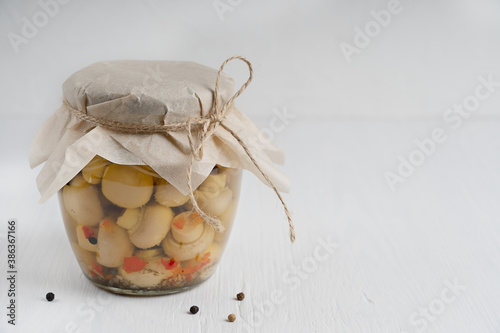 Single glass jar full of homemade organic marinated or fermented champignon mushrooms decorated with paper on white wooden background with black pepper. Image with copy space, horizontal orientation