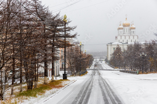 Snow-covered city street during a snowfall. Cold snowy weather. Slippery road, sleet and ice. View of the beautiful cathedral. City of Magadan, Magadan region, Siberia, Far East Russia. Mid October.