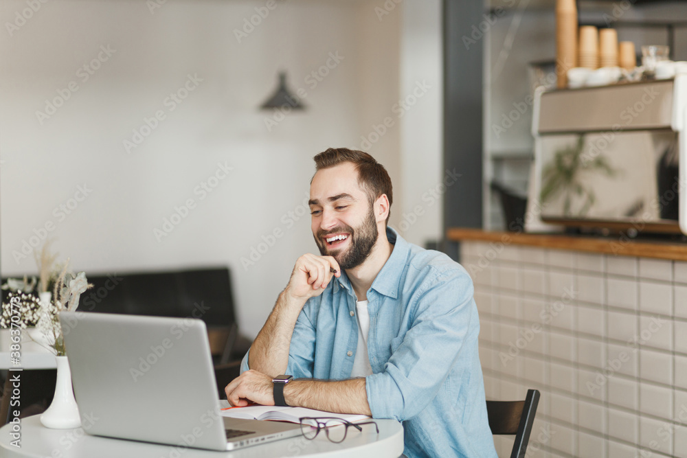 Smiling young man sitting alone at table in coffee shop cafe working studying on laptop pc computer hold pen relaxing in restaurant during free time indoors. Freelance mobile office business concept.