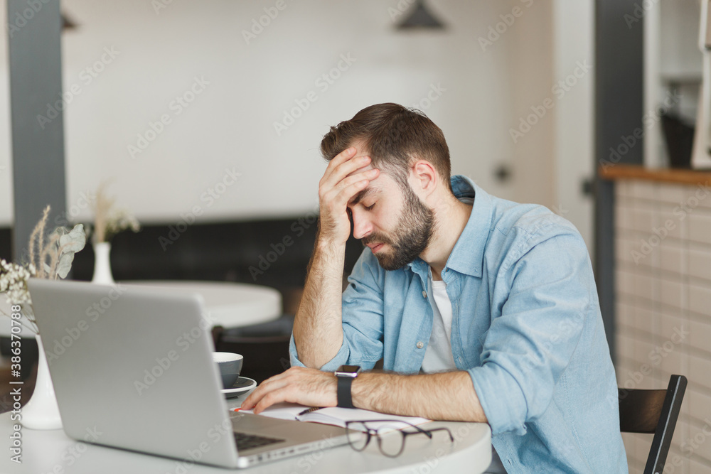 Sick tired exhausted young man sitting alone at table in coffee shop cafe restaurant indoor working or studying on laptop pc computer put hand on head. Freelance mobile office business concept. Sick tired exhausted young man sitting alone at table in coffee shop cafe restaurant indoor working or studying on laptop pc computer put hand on head. Freelance mobile office business concept.