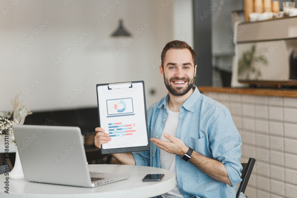 Smiling young man sit at table in coffee shop cafe restaurant indoors working or studying on laptop pc computer pointing hand clipboard with papers document. Freelance mobile office business concept. Smiling young man sit at table in coffee shop cafe restaurant indoors working or studying on laptop pc computer pointing hand clipboard with papers document. Freelance mobile office business concept.