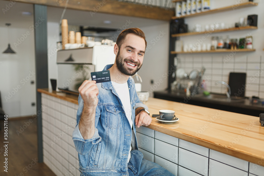 Smiling young man in denim jacket sit alone in coffee shop cafe with cup of coffee tea hold credit bank card relaxing in restaurant during free time indoors. Freelance mobile office business concept.
