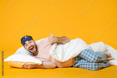 Full length of surprised excited cheerful man in pajamas home wear sleep mask lying showing thumb up isolated on bright yellow colour background studio portrait. Relax good mood lifestyle concept.