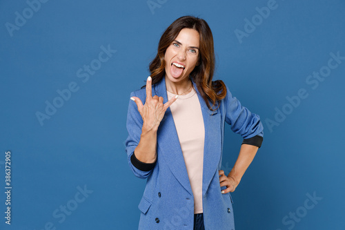 Cheerful funny crazy young brunette woman 20s wearing basic jacket showing horns up gesture, depicting heavy metal rock sign looking camera isolated on bright blue colour background studio portrait.